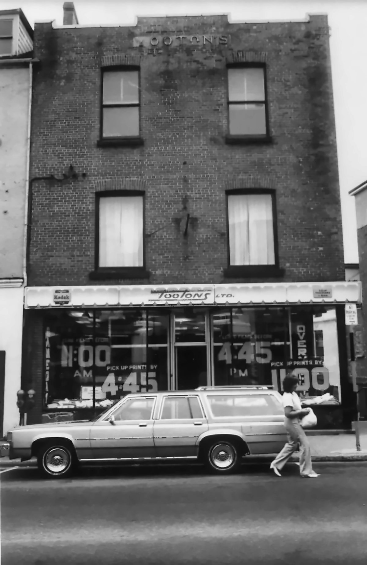 Black-and-white photograph of the Tooton&rsquo;s Ltd. storefront, a three-storey brick building with Kodak signs and &ldquo;Pick up prints by 4:45&rdquo; in the windows
